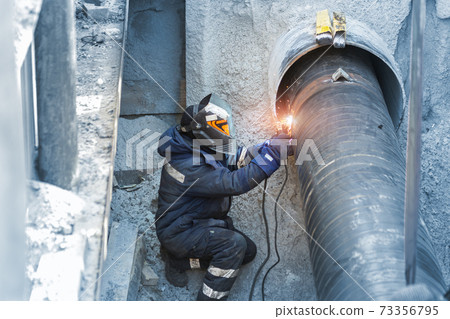Professional welder welding water or gas steel pipeline in protective trench and mask at construction site pit. City underground utilitites renewal replacement. Emergency pipe break leak elimination 73356795