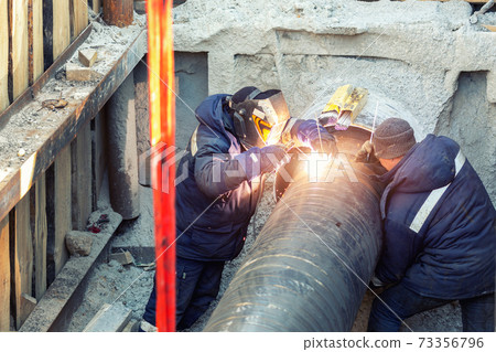 Professional welder welding water or gas steel pipeline in protective trench and mask at construction site pit. City underground utilitites renewal replacement. Emergency pipe break leak elimination Professional welder welding water or gas steel pipeline in protective trench and mask at construction site pit. City underground utilitites renewal replacement. Emergency pipe break leak elimination 73356796