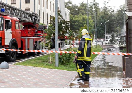 Firemen un uniform standing behind fire tape and many fire engine trucks with ladder at accident in highrise tower residential or office building in city center. Emergency rescue at disaster Firemen un uniform standing behind fire tape and many fire engine trucks with ladder at accident in highrise tower residential or office building in city center. Emergency rescue at disaster 73356799