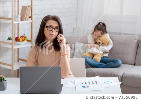 Modern working mom doing online job, her neglected daughter hugging teddy bear, feeling lonely and sad at home Modern working mom doing online job, her neglected daughter hugging teddy bear, feeling lonely and sad at home 73356983