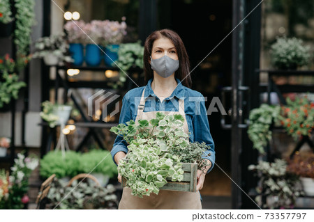 Floristics, business, decorating and gardening. Friendly female in apron and protective mask carries box of plants Floristics, business, decorating and gardening. Friendly female in apron and protective mask carries box of plants 73357797