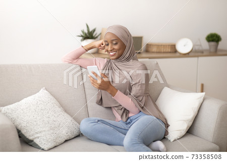 Millennial Muslim lady in hijab using mobile phone at home, messaging or browsing social media, sitting on couch indoors 73358508