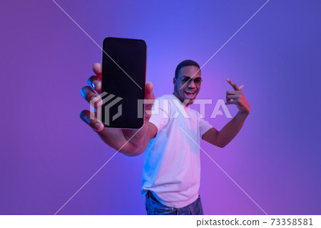Mobile Offer. Excited African Guy Demonstrating Smartphone With Black Screen, Neon Lighting 73358581