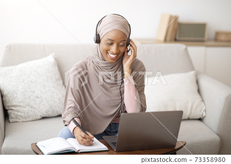 Joyful African American woman in hijab and headphones taking notes during online business meeting on laptop at home 73358980