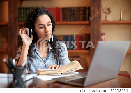Lady sitting at desk, wearing headset, using computer, reading book Lady sitting at desk, wearing headset, using computer, reading book 73358984