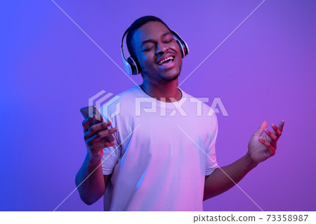 Black guy in headphones listening music on smartphone and singing, neon lighting Black guy in headphones listening music on smartphone and singing, neon lighting 73358987