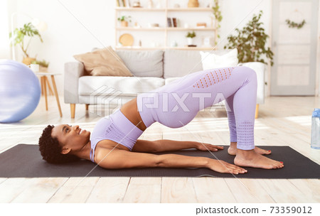 Sporty black woman doing exercises on yoga mat, standing in half bridge pose indoors, side view Sporty black woman doing exercises on yoga mat, standing in half bridge pose indoors, side view 73359012