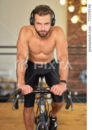 Athletic young man in headphones riding stationery bicycle Athletic young man in headphones riding stationery bicycle 73359536