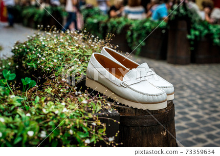 White leather moccasins. Photo on the street next to the flowers. Trendy white loafers. Fashionable women's leather shoes. White leather moccasins. Photo on the street next to the flowers. Trendy white loafers. Fashionable women's leather shoes. 73359634