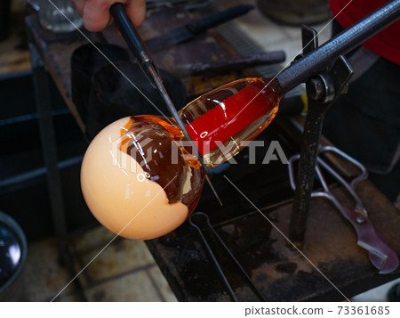 Glassblower is blowing into melted glass and add white layer. 73361685