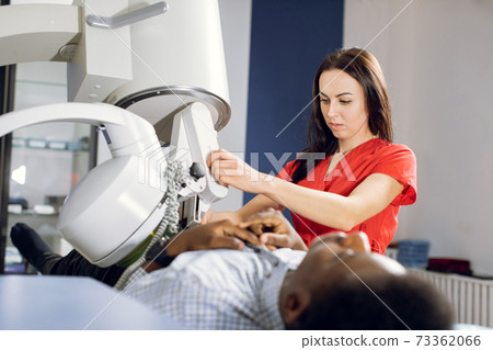 Procedure of extracorporeal shock wave lithotripsy in modern urology medical center. Close up of pretty Caucasian woman doctor providing stone treatment for black male patient using lithotripter 73362066