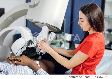 Close up side view of high-skilled female doctor in red uniform, performing lithotripsy, using modern lithotripter to break up kidney stones of male African patient with high-intensity acoustic pulse Close up side view of high-skilled female doctor in red uniform, performing lithotripsy, using modern lithotripter to break up kidney stones of male African patient with high-intensity acoustic pulse 73362069