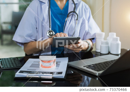 Concentrated dentist sitting at table with jaw samples tooth model and working with tablet and laptop in dental office professional dental clinic.. 73370466