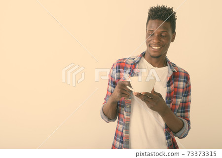 Studio shot of young happy black African man smiling while looking at coffee cup against white Studio shot of young happy black African man smiling while looking at coffee cup against white 73373315