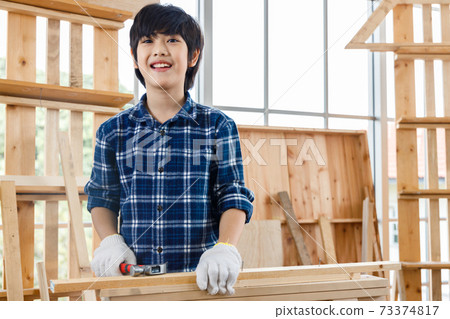 Portrait of happy Asian boy standing in carpentry workshop and holding hammer at home. Portrait of happy Asian boy standing in carpentry workshop and holding hammer at home. 73374817