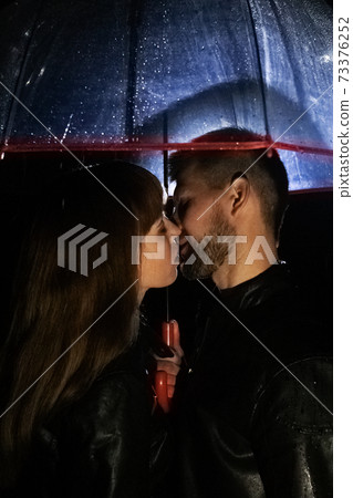 A couple in love under an umbrella kissing, and rain is falling on them. Night photography of people. The concept of protecting relationships and long love for a couple. A couple in love under an umbrella kissing, and rain is falling on them. Night photography of people. The concept of protecting relationships and long love for a couple. 73376252