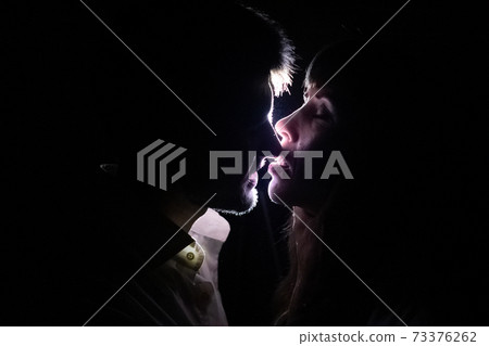 A couple of lovers kiss at night against the light. Against a black background, rays are visible passing near the faces and creating the effect of silhouettes of people. 73376262