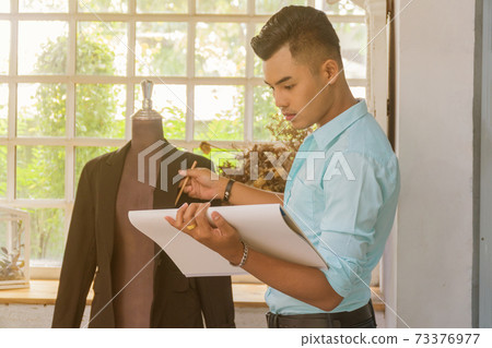 Asian young fashion designer holding a book standing beside the mannequins. Asian young fashion designer holding a book standing beside the mannequins. 73376977