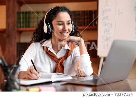 Woman sitting at table, using laptop and writing in notebook 73377154