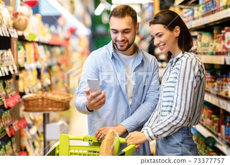 Young couple with cart and smartphone shopping in supermarket Young couple with cart and smartphone shopping in supermarket 73377757