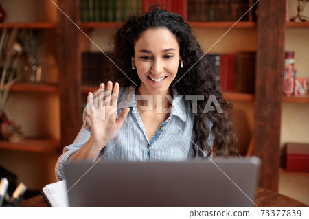 Woman sitting at desk, using laptop and waving to webcam 73377839