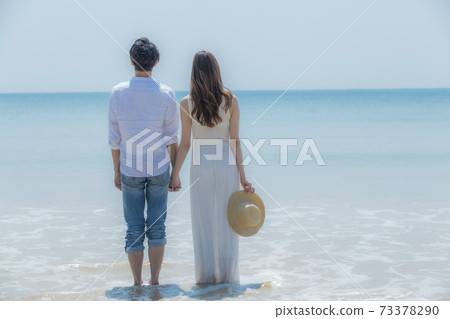 The back of a couple holding hands at the edge of a beach 73378290