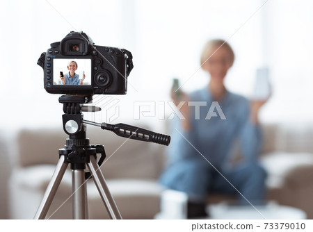 Young Caucasian woman recording video content for her tech blog at home, selective focus on camera with preview screen 73379010