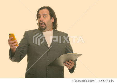 Fat Caucasian businessman looking shocked while using mobile phone and holding clipboard Fat Caucasian businessman looking shocked while using mobile phone and holding clipboard 73382419