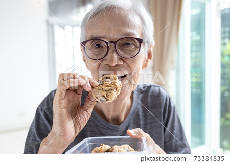 Happy asian senior woman eating chocolate chip cookie or tasting sweet cookie,natural tasty food,delicious snack,old elderly holding a box of cashew nut biscuits in her hand,breakfast in the morning Happy asian senior woman eating chocolate chip cookie or tasting sweet cookie,natural tasty food,delicious snack,old elderly holding a box of cashew nut biscuits in her hand,breakfast in the morning 73384835