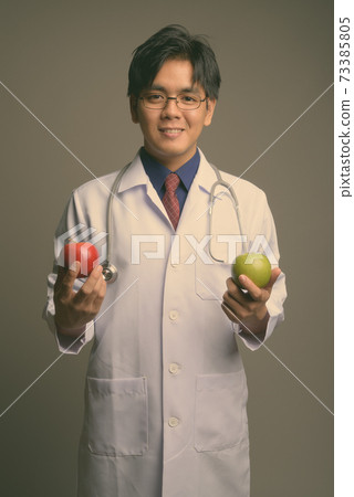 Young handsome Asian man doctor wearing eyeglasses against gray background Young handsome Asian man doctor wearing eyeglasses against gray background 73385805