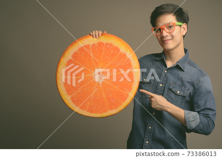 Young handsome Asian man holding orange pillow against gray background 73386013