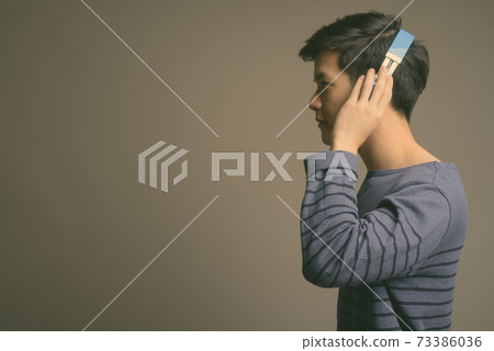 Young handsome Asian man listening to music against gray background Young handsome Asian man listening to music against gray background 73386036