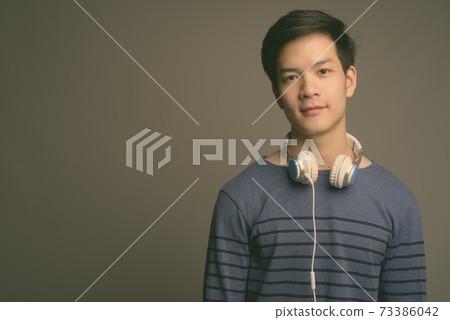 Young handsome Asian man wearing headphones against gray background 73386042
