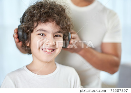 Happy kid. Portrait of joyful latin boy smiling at camera while spending time at home together with his father. Dad putting on headphones on his child in the background Happy kid. Portrait of joyful latin boy smiling at camera while spending time at home together with his father. Dad putting on headphones on his child in the background 73387897