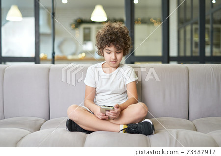 Modern childhood. Cute latin boy sitting on a sofa with smartphone, texting message or playing game while spending time at home Modern childhood. Cute latin boy sitting on a sofa with smartphone, texting message or playing game while spending time at home 73387912