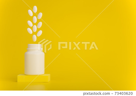 Opened medication bottle on podium with white oval pills jumping out against yellow background with copy space 73403620