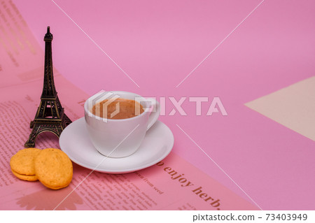 Coffee in a white Cup with a saucer and colorful macaroons next to the Eiffel tower statuette on a pink background and wrapping paper with newspaper decor. layout Coffee in a white Cup with a saucer and colorful macaroons next to the Eiffel tower statuette on a pink background and wrapping paper with newspaper decor. layout 73403949