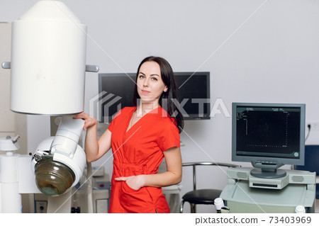 Close up front view of high-skilled female doctor in red uniform, standing near the modern machine for non-invasive extracorporeal shock wave lithotripsy to break up kidney or ureter stones 73403969