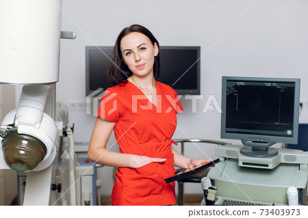 Close up portrait of young caucasian woman doctor in red uniform, holding digital tablet pc, posing near modern ultrasonic lithotripter machine in urology medical center 73403973