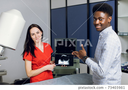Close up portrait of smiling African man, posing to camera, while visiting his female doctor in modern clinic, holding digital tablet with ultrasound scan. Focus on the woman doctor Close up portrait of smiling African man, posing to camera, while visiting his female doctor in modern clinic, holding digital tablet with ultrasound scan. Focus on the woman doctor 73403981