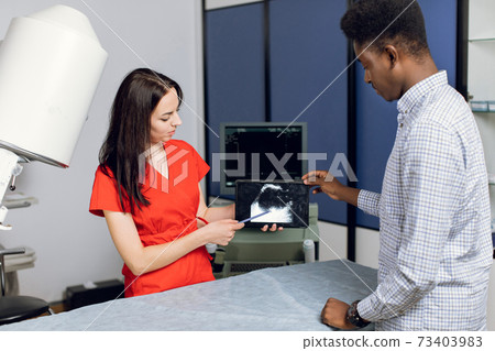 Professional woman ultrasound specialist, holding ipad pc with ultrasound scan image and explaining ways of treatment kidney stones for her young male African American patient 73403983