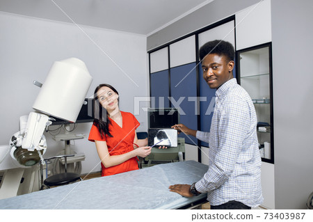 Happy doctor and patient in modern urology clinic, celebrating successful treatment. Young female doctor and her male african patient, holding tablet pc with ultrasound scan, smiling to camera 73403987