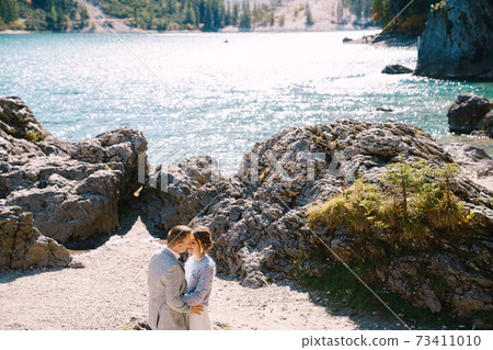 Bride and groom stand against the backdrop of stones overlooking Lago di Braies in Italy. Destination wedding in Europe, on Braies lake. Loving newlyweds walk against the backdrop of amazing nature. 73411010