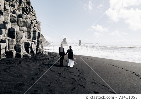 Destination Iceland wedding. A wedding couple walks along the sandy black beach of Vik, near the basalt rock, in the form of pillars. The bride and groom hold hands and walk along the shore. Destination Iceland wedding. A wedding couple walks along the sandy black beach of Vik, near the basalt rock, in the form of pillars. The bride and groom hold hands and walk along the shore. 73411055
