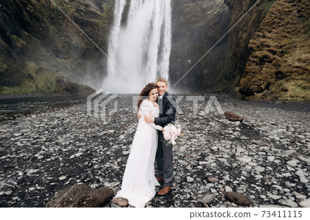 The groom hugs the bride, it is snowing. Wedding couple near Skogafoss waterfall. Destination Iceland wedding. The groom hugs the bride, it is snowing. Wedding couple near Skogafoss waterfall. Destination Iceland wedding. 73411115