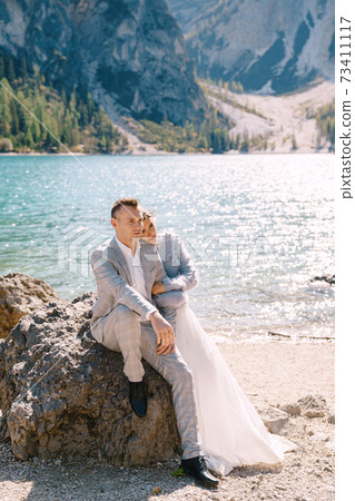 Bride and groom sit embracing against background of stones at the Lago di Braies in Italy. Destination wedding in Europe, on Braies lake. Loving newlyweds walk against the backdrop of amazing nature Bride and groom sit embracing against background of stones at the Lago di Braies in Italy. Destination wedding in Europe, on Braies lake. Loving newlyweds walk against the backdrop of amazing nature 73411117