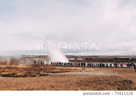 Tourists look at the eruption of a geyser. Geyser Valley in southwest of Iceland. The famous tourist attraction Geysir. Geothermal zone Haukadalur. Strokkur geyser on the slopes of Laugarfjall hill. 73411129