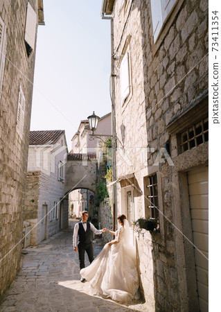 Fine-art wedding photo in Montenegro, Perast. The bride and groom on the street of the old city, among the old houses. Fine-art wedding photo in Montenegro, Perast. The bride and groom on the street of the old city, among the old houses. 73411354