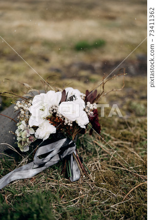 bridal bouquet of white roses, lily, burgundy cloves, protea, dried branches, brunia and leucadendron with grey and black ribbons near the dried grass 73411362