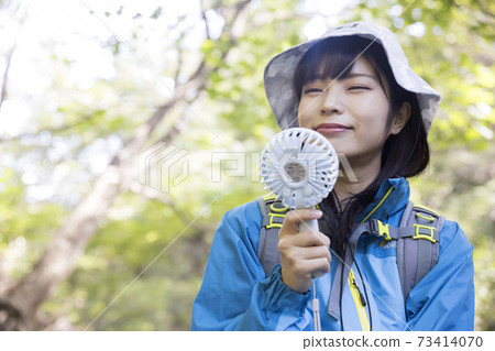 徒步旅行期間用便攜式風扇冷卻的年輕女子徒步旅行圖像 徒步旅行期間用便攜式風扇冷卻的年輕女子徒步旅行圖像 73414070